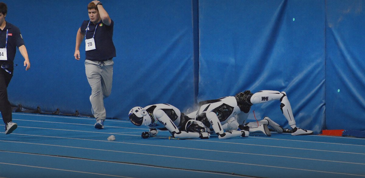 Two humanoid robots collapsed against the padded blue crash wall at the end of a sprint track, with human handlers running in from the left.
